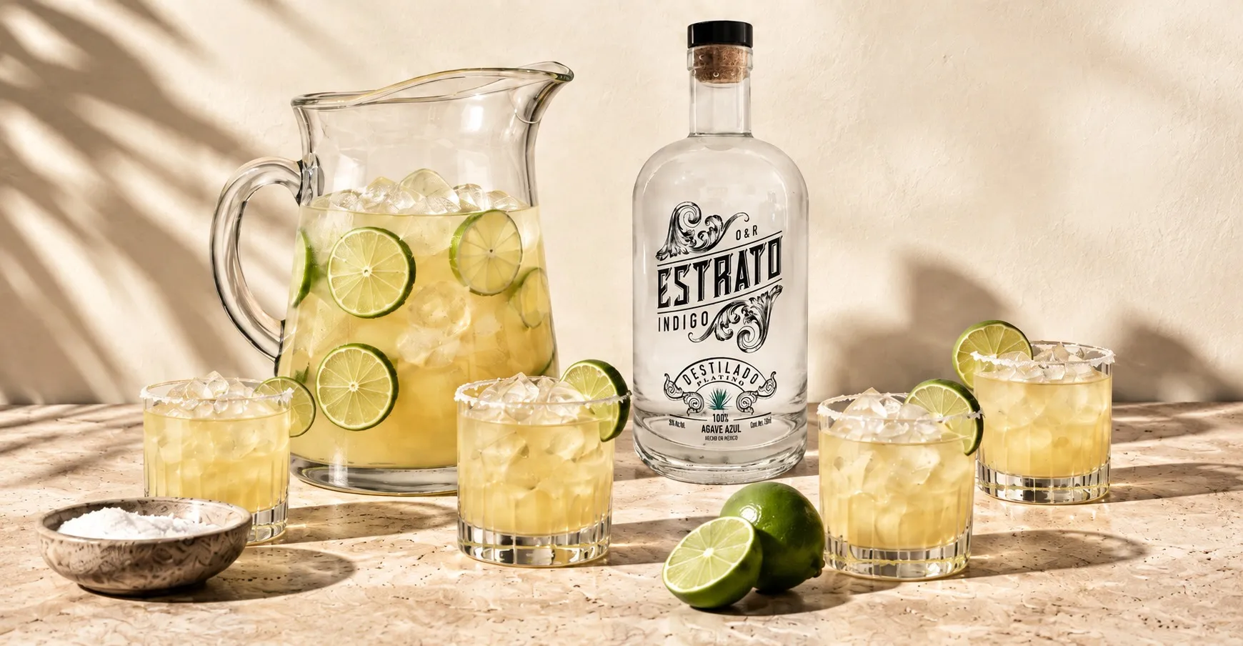 A glass pitcher of margarita with lime wheels beside a bottle of Estrato and four salt-rimmed rocks glasses on a sunlit stone table